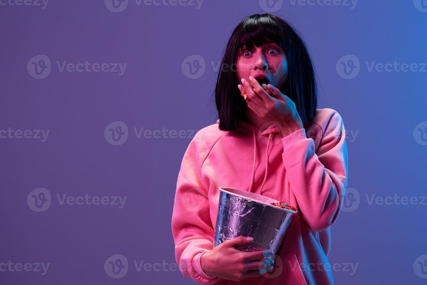 Scared shocked awesome brunet woman in pink hoodie eat popcorn look aside posing isolated in blue violet color light studio background. Neon party Cyberpunk Cinema concept. Copy space photo