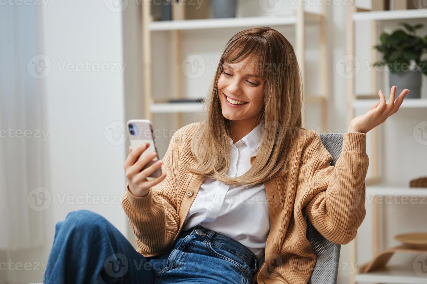 emocionado alegre joven rubia dama en calentar suéter hacer vídeo llamada saludar en línea amigo sentado en Sillón a hogar interior. pausa desde trabajar, tomar un romper, social medios de comunicación en gratis hora concepto. Copiar espacio foto