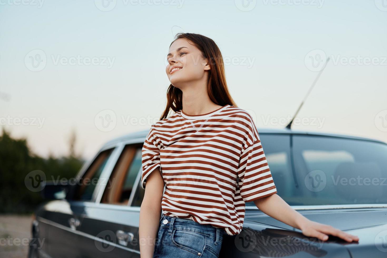 A young woman in a striped T-shirt and jeans is relaxing from driving, leaning on her car beside the road and smiling at the nature and the sunset photo
