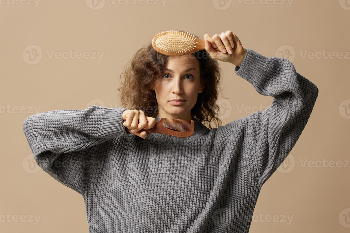 encantador contento Rizado hermosa hembra en gris casual suéter sostener cepillo para el pelo peine en frente de cámara posando aislado en terminado beige pastel antecedentes. problemático revoltoso dañado pelo concepto. Copiar espacio foto