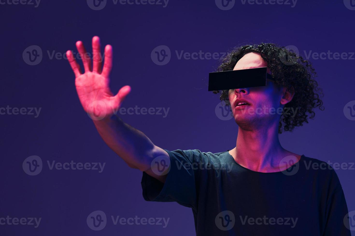 Excited stylish tanned curly man in black t-shirt eyewear touches invisible object posing isolated on color pink blue background. Cool fashion offer. Virtual Reality New Collection concept. Copy space photo