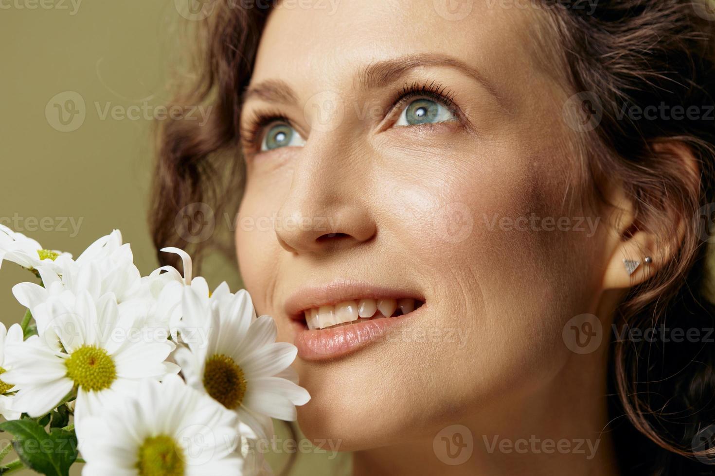 Close up portrait of smiling curly female in linen casual shirt hold chamomiles flowers near face looks up posing isolated on over olive green pastel background. Nature is beauty concept. Copy space photo