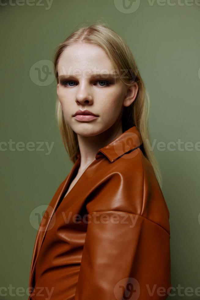 Handsome pretty blonde stylish young lady in brown palette leather dress serious looking over her shoulder at camera posing isolated on over olive green studio wall background. Fashion portrait banner photo