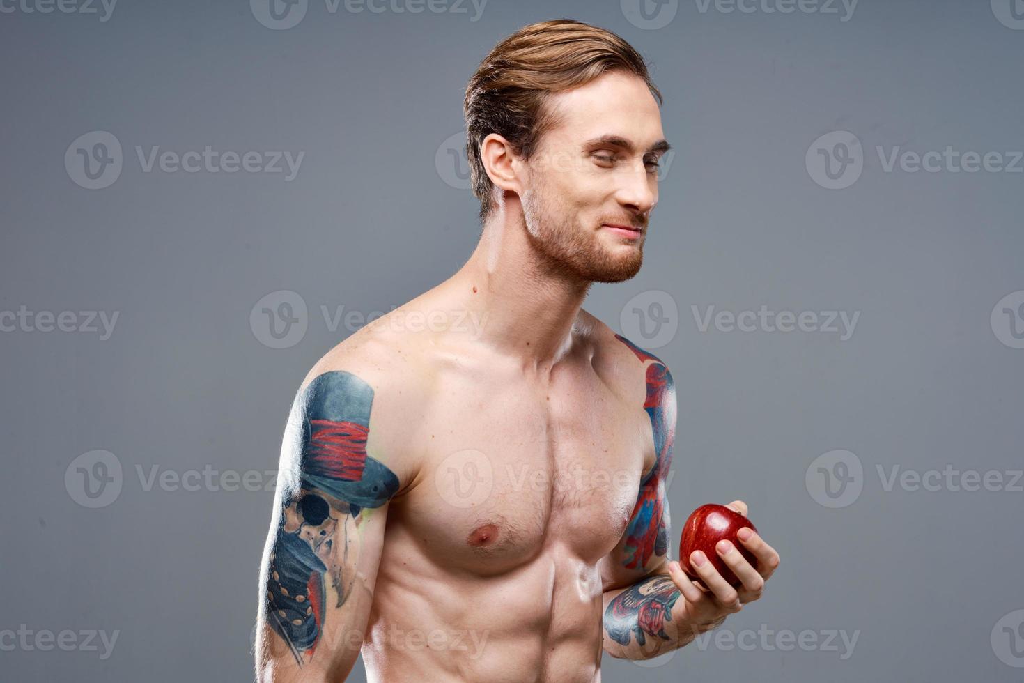 sexy athlete with pumped up muscles eats an apple on a gray background and a tattoo on his arm photo