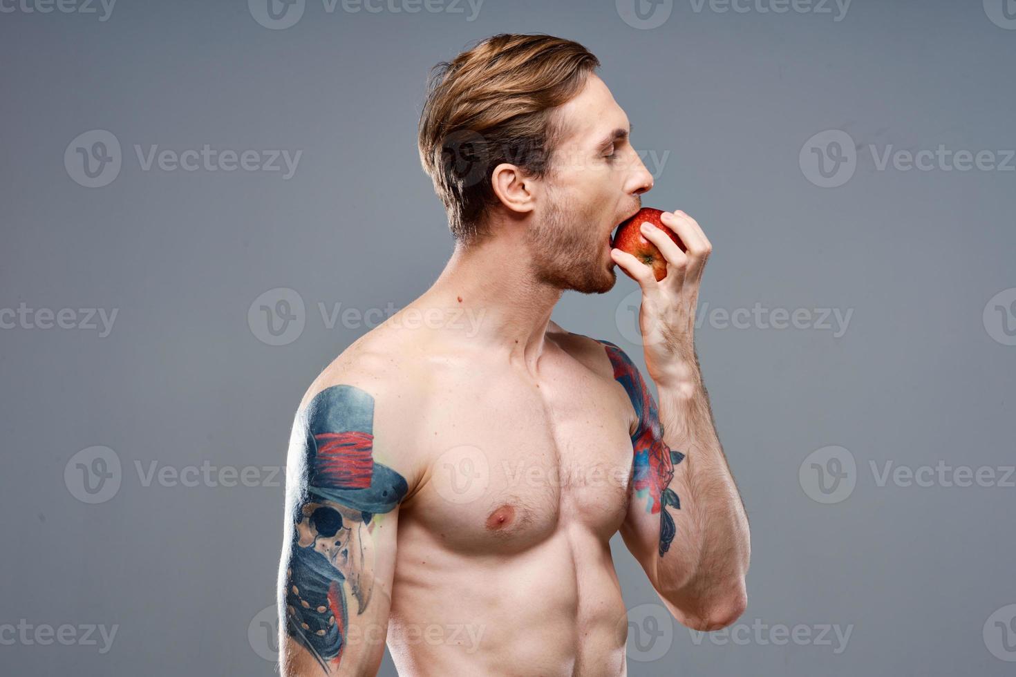 sexy athlete with pumped up muscles eats an apple on a gray background and a tattoo photo