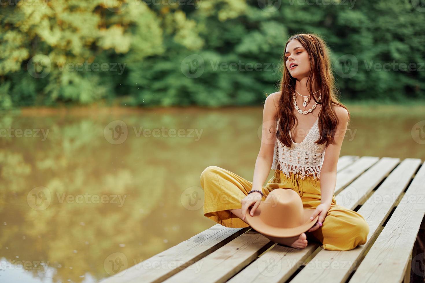 A young hippie woman sits on a lake bridge wearing stylish eco clothes and smiling photo