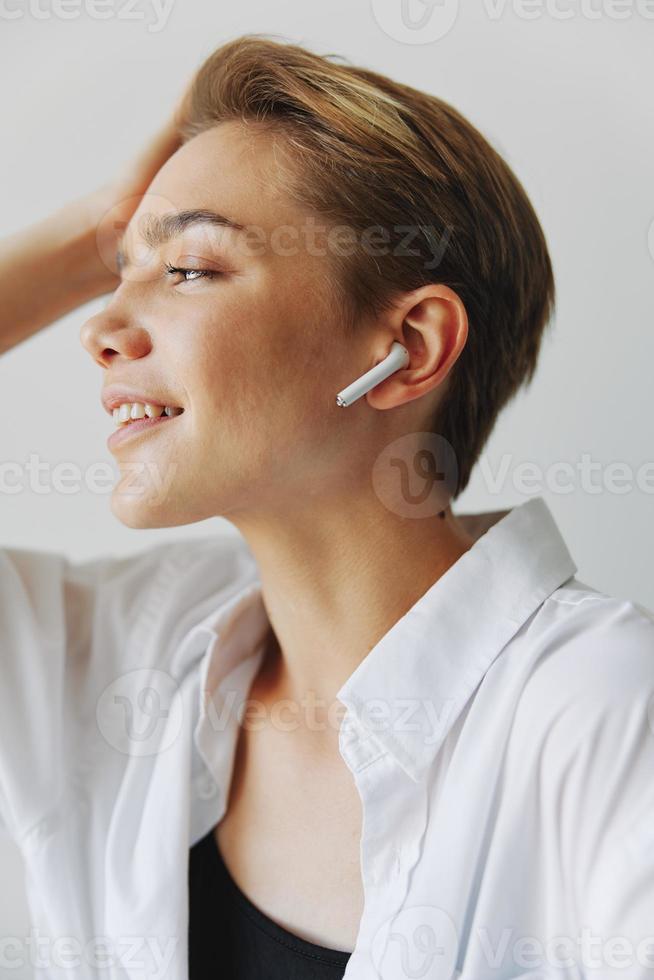 joven mujer adolescente escuchando a música con estéril auriculares y bailando hogar, sonriendo con dientes con un corto Corte de pelo en un blanco camisa en un blanco antecedentes. niña natural poses con No filtros foto