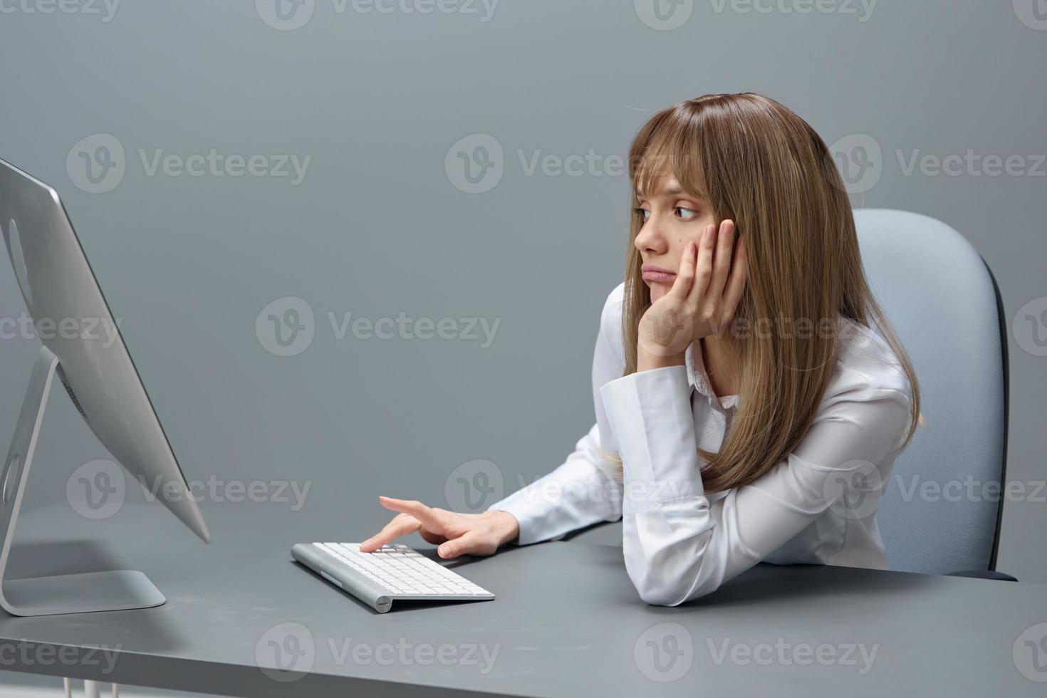 Bored pretty blonde businesswoman worker freelancer using desktop computer sitting at workplace in gray modern office. Remote Job, Technology And Career Profession Concept. Copy space photo