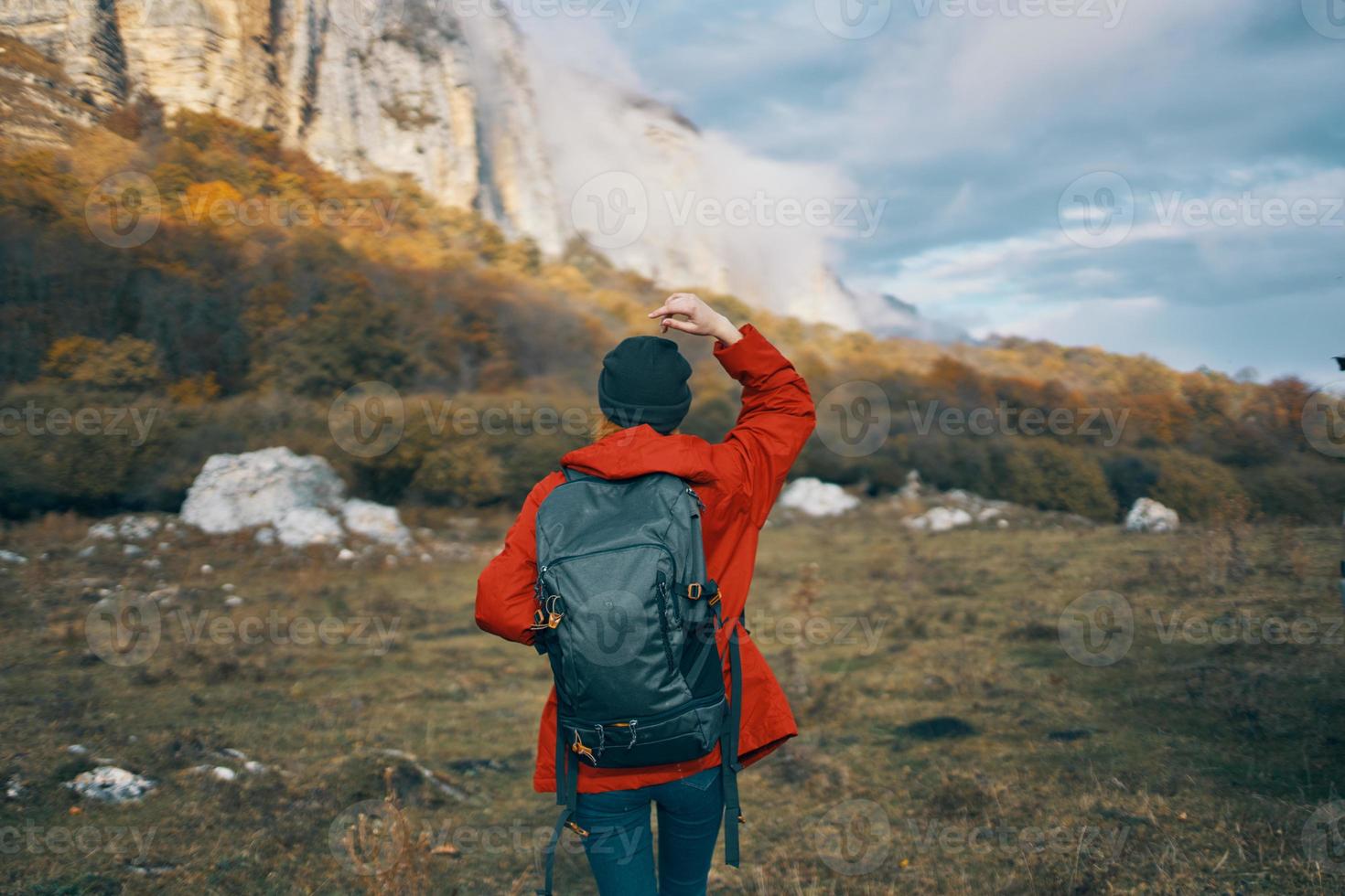 woman in a gray hat red jacket with a backpack gestures with her hands on nature in the mountains photo