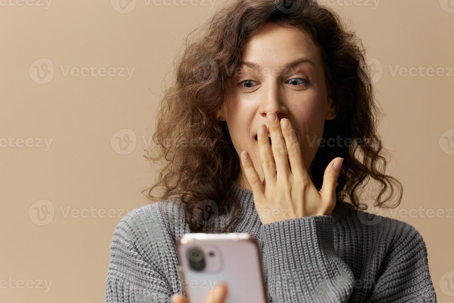 Shocked curly female in gray casual sweater read gossip info in phone close mouth with hand posing isolated on over beige background. Social media, network, distance communication concept. Copy space photo
