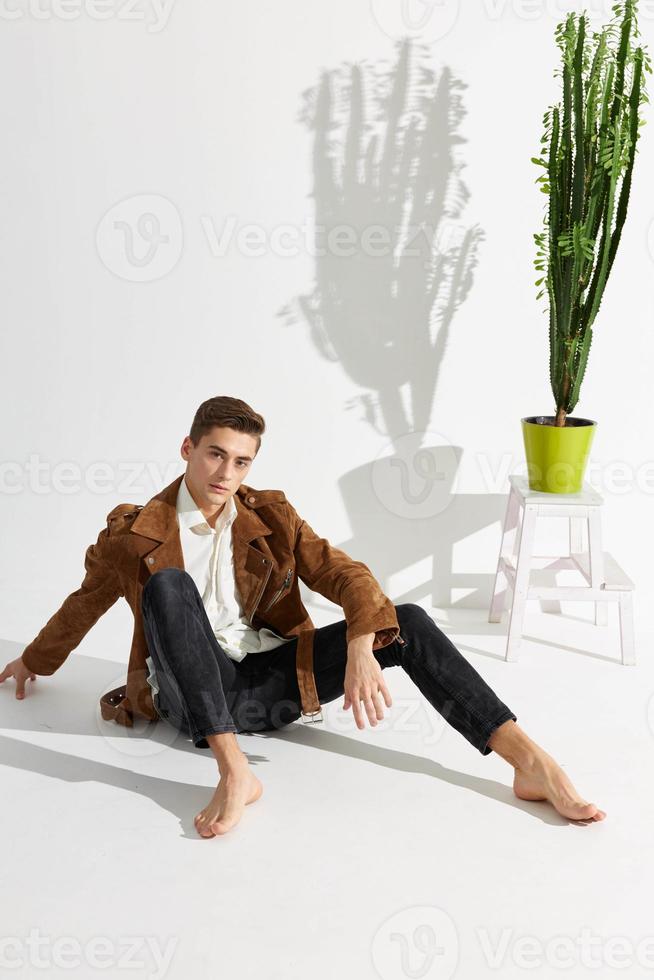 A handsome young man sits on the floor in a jacket and black trousers and a flower in a pot on a chair photo