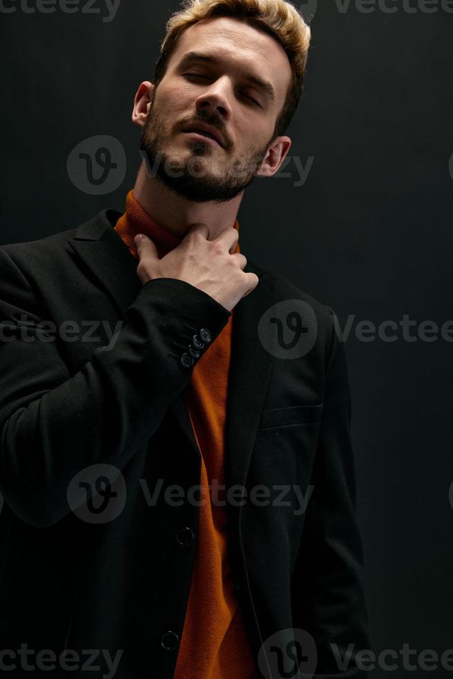 a blond guy in an orange sweater and a jacket holds his hand near his neck photo