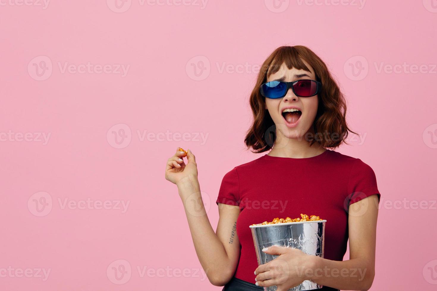 Overjoyed happy cute redhead lady in red t-shirt 3D glasses with popcorn watch movie posing isolated on over pink studio background. Copy space Banner. Fashion Cinema concept. Entertainment offer photo