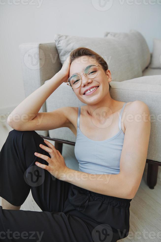 Teenage girl sitting on the floor at home smiling in home clothes and glasses with a short haircut, lifestyle without filters, free copy space photo