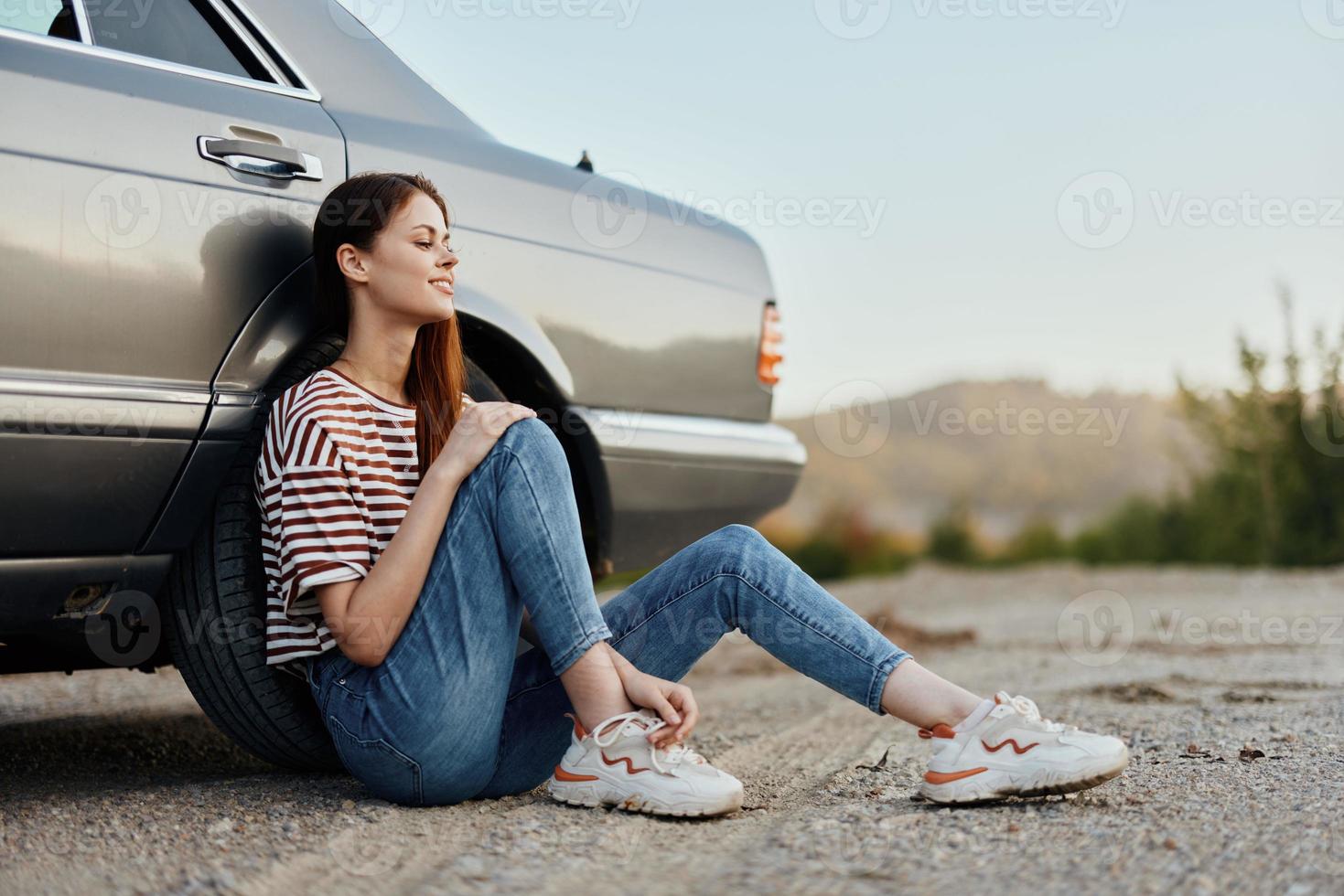 A young woman sits on the ground near her car on the side of the road and looks at the sunset photo