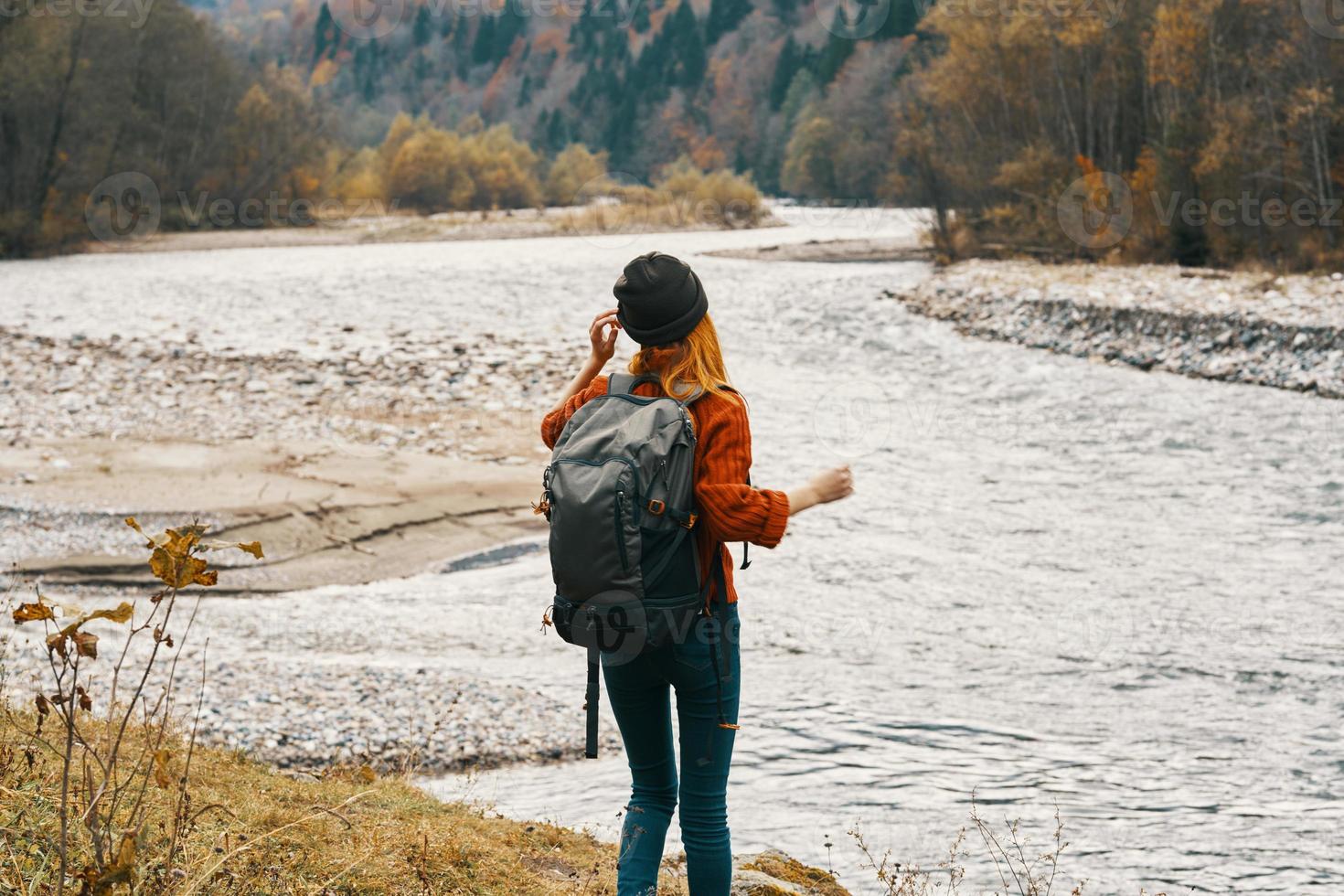 a traveler in a sweater hat with a backpack gestures with her hands and relaxes on the river bank in the mountains photo