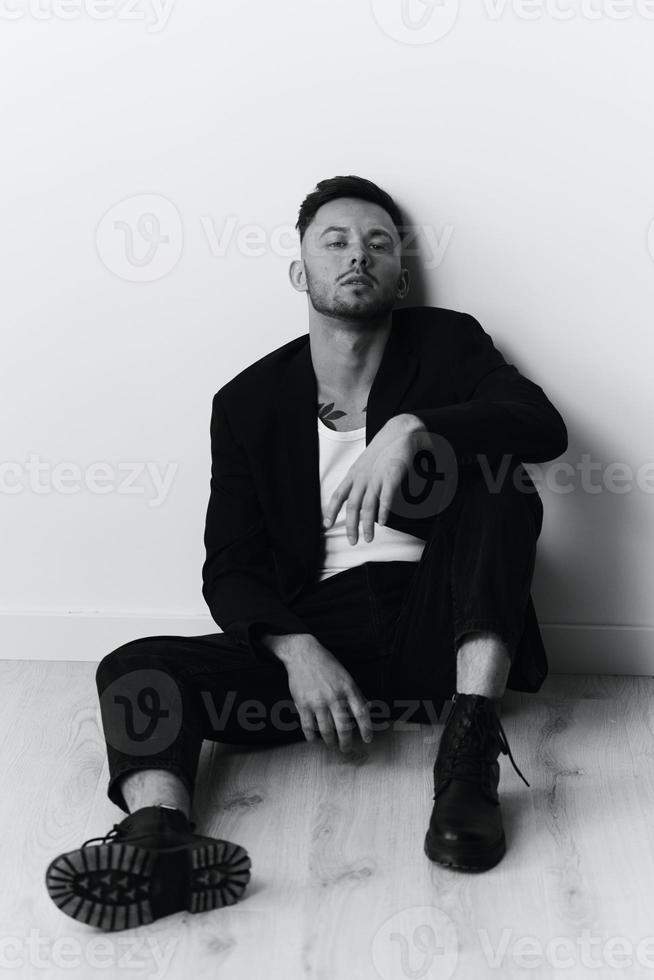 Modelling snapshots. Serious self-confident attractive handsome man in black jacket sitting on floor posing in white studio background. Black and White concept. Copy space photo