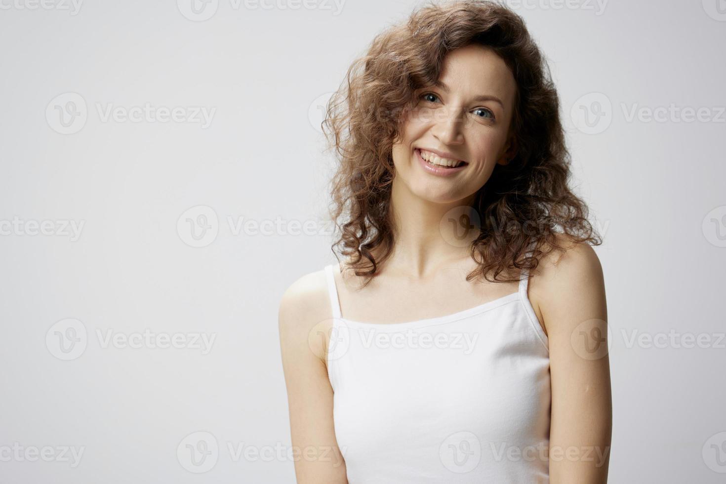 linda contento disfruté Rizado hermosa mujer en básico blanco camiseta sonrisas a cámara posando aislado en terminado blanco antecedentes. personas emociones estilo de vida concepto. Copiar espacio foto