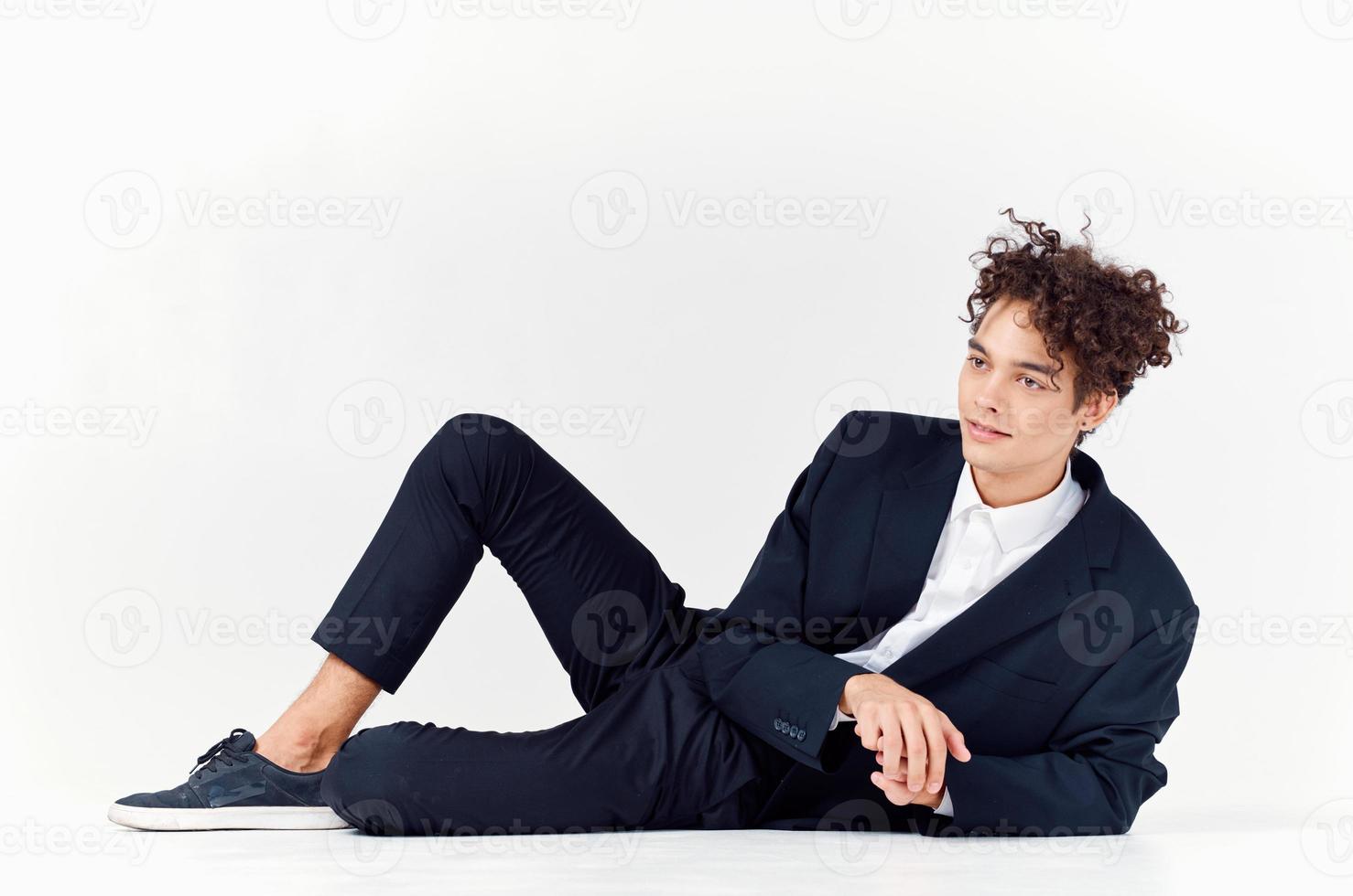 a man with curly hair lies in a suit on the floor in a bright room and sneakers style photo