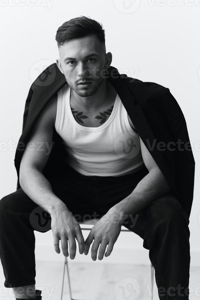 Modelling snapshots. Serious self-confident attractive handsome man in black jacket sitting on chair posing in white studio background. Black and White concept. Copy space photo