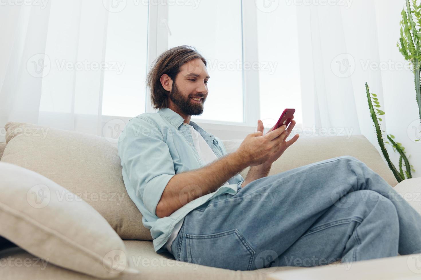 A man with a beard sits on the couch during the day and smiles at home and looks at his phone relaxing on his day off, life online on his phone texting with friends photo