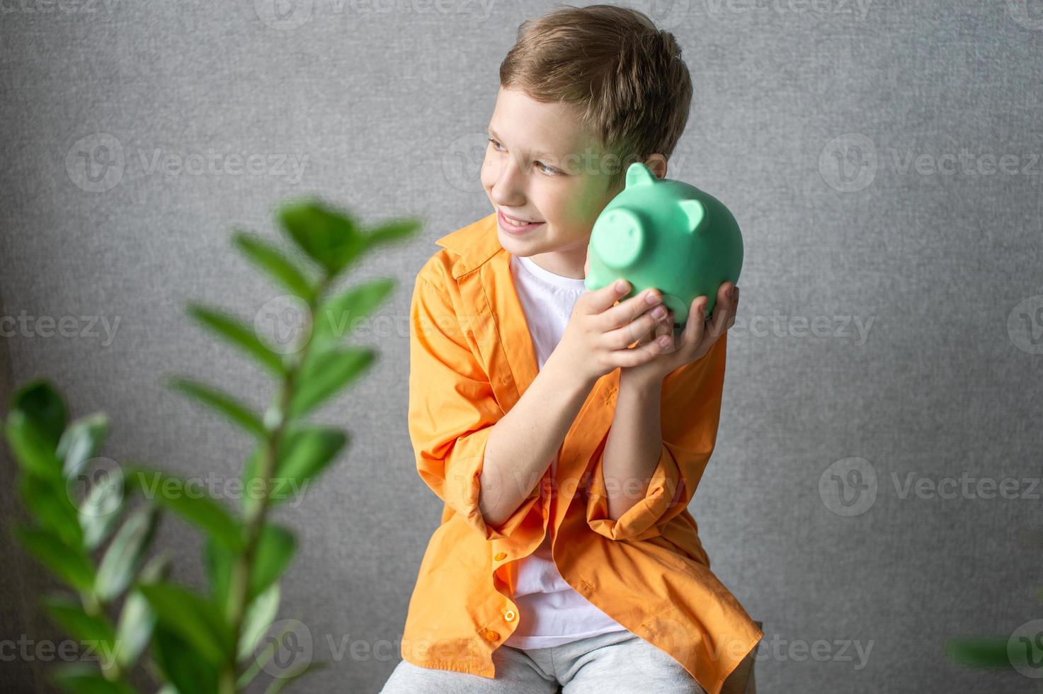 A cute boy in a shirt shakes a piggy bank and listens to the clinking of coins photo