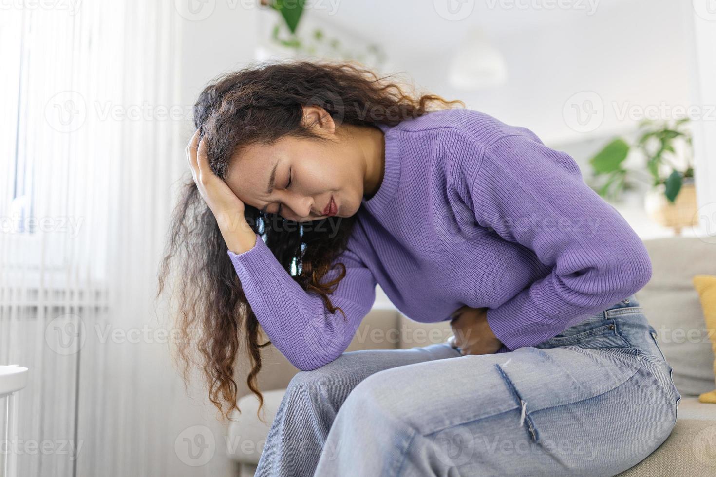 Asian woman in painful expression holding hands against belly suffering menstrual period pain, lying sad on home bed, having tummy cramp in female health concept photo