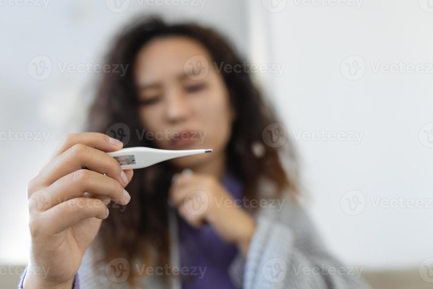 Asian woman is having flu and she is using thermometer. Sick with a rhinitis womanwith hijab has dripping nose. Woman being sick having flu lying on sofa looking at temperature on thermometer. photo