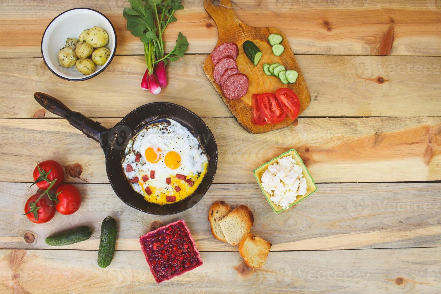 Different food scrambled eggs in frying pan, boiled potatoes, curd, croutons, radishes, cucumbers, tomatoes, smoked sausage, croutons, mint, sorrel on wooden table photo