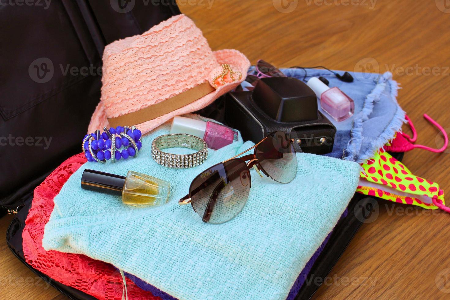 Open the suitcase with tourist things women's hat, swimsuit, camera, denim shorts, dresses, sunglasses, perfumes, nail polish, bracelets. photo