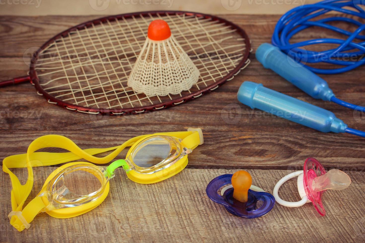 Pacifier and sports equipment the birdie is on the racket, skipping rope, swimming goggles on wooden background. Concept of sports to be engaged with early childhood. Toned image. photo