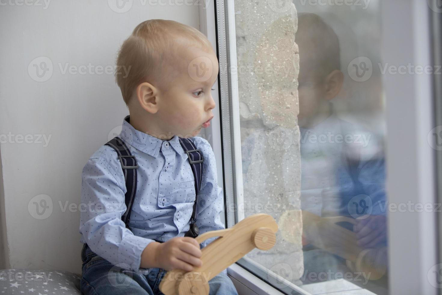 A small child looks out the window. The kid with a toy sits on a window sill. photo