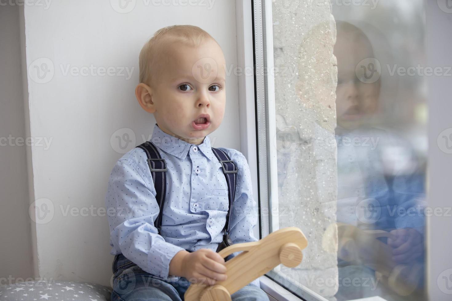 Little baby near the window. A kid with a wooden typewriter is sitting on a windowsill. photo