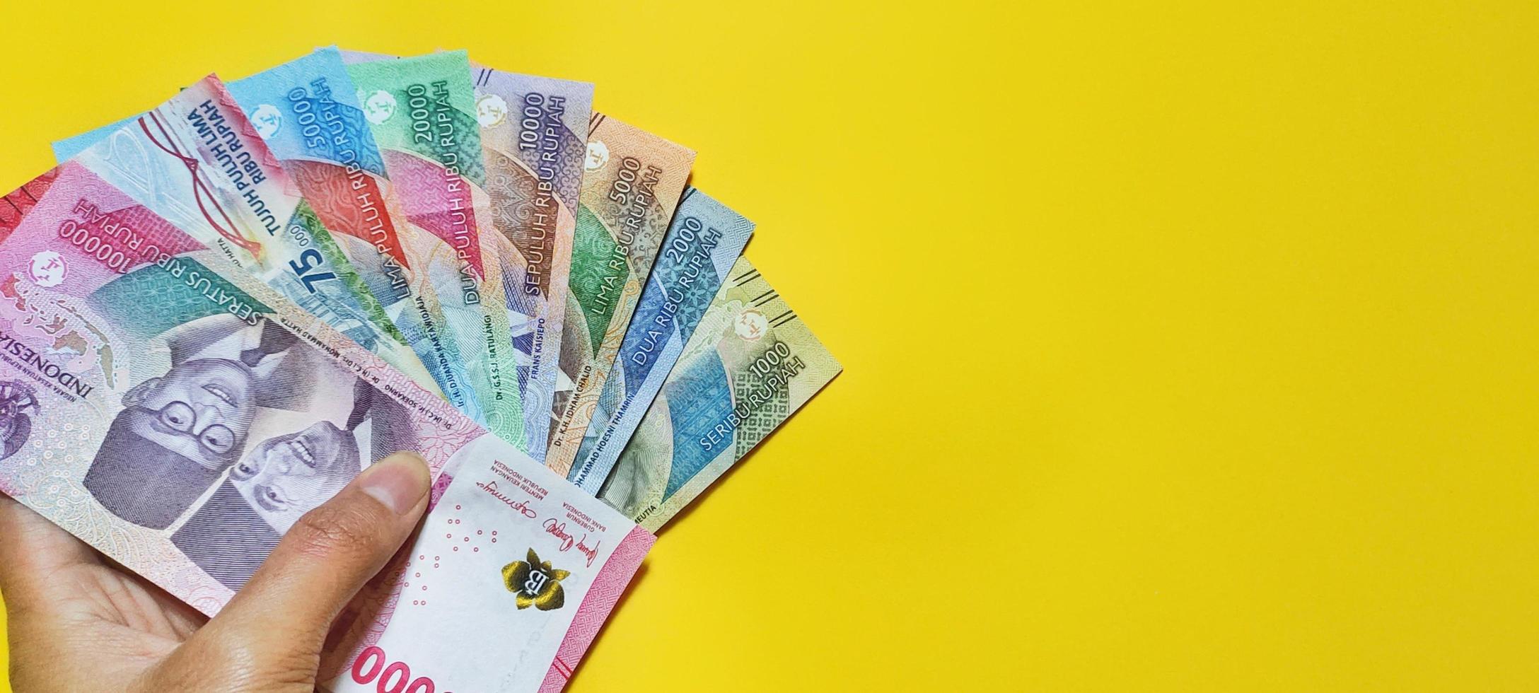 Man holding new banknotes issued in 2022 from Rp.1,000 to Rp.100,000. Indonesian rupiah currency, Tunjangan Hari Raya concept isolated on yellow background. Negative space photo