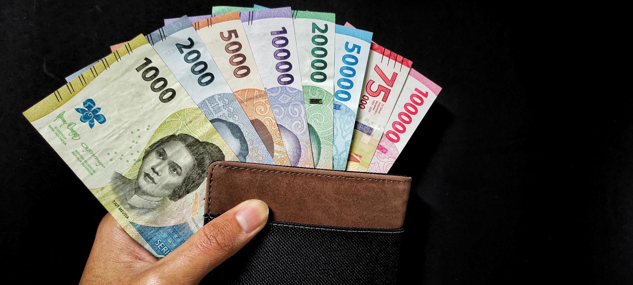 hombre participación nuevo billetes emitido en 2022 desde 1000 rupias a 100.000 rupias y un marrón billetera. indonesio rupia moneda concepto aislado en negro antecedentes. negativo espacio foto