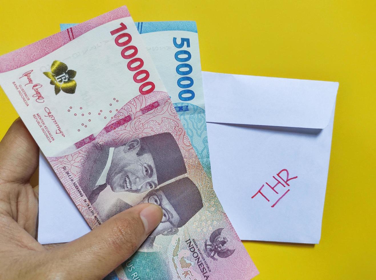 Man holding a white envelope written of THR and new Indonesian banknotes, usually Tunjangan Hari Raya or called THR are given to employees ahead of Eid. Isolated on black background and top view photo