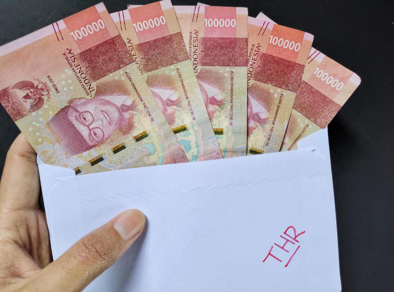 The man holds a white envelope with the words THR containing Indonesian banknotes of IDR 100,000. Usually the Hari Raya Allowance or known as THR is given to employees ahead of Eid. Top view photo