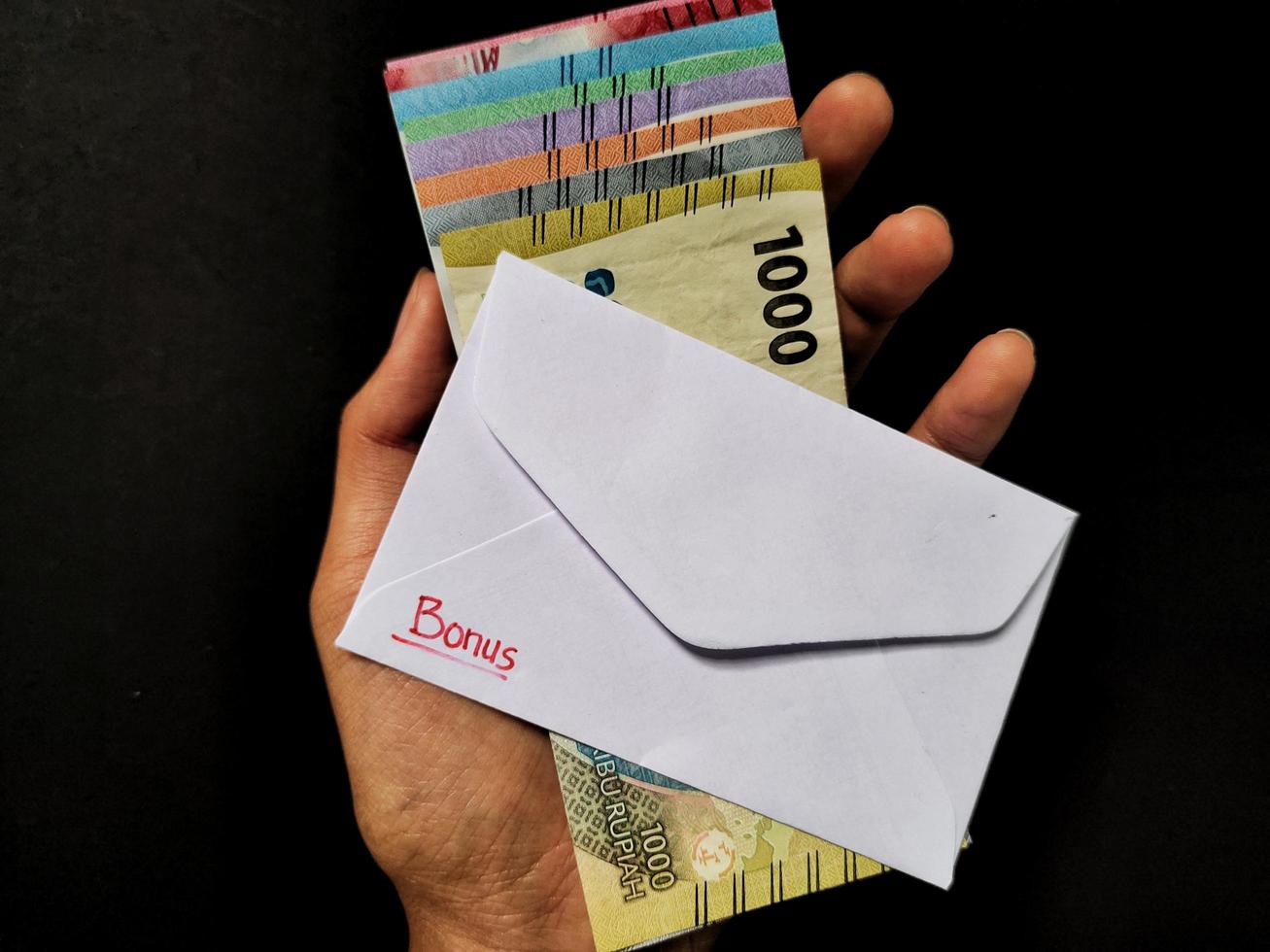 Man holding a white envelope written bonus and new Indonesian banknotes. Usually the Holiday Allowance or known as THR is given to employees ahead of Eid. Top view photo