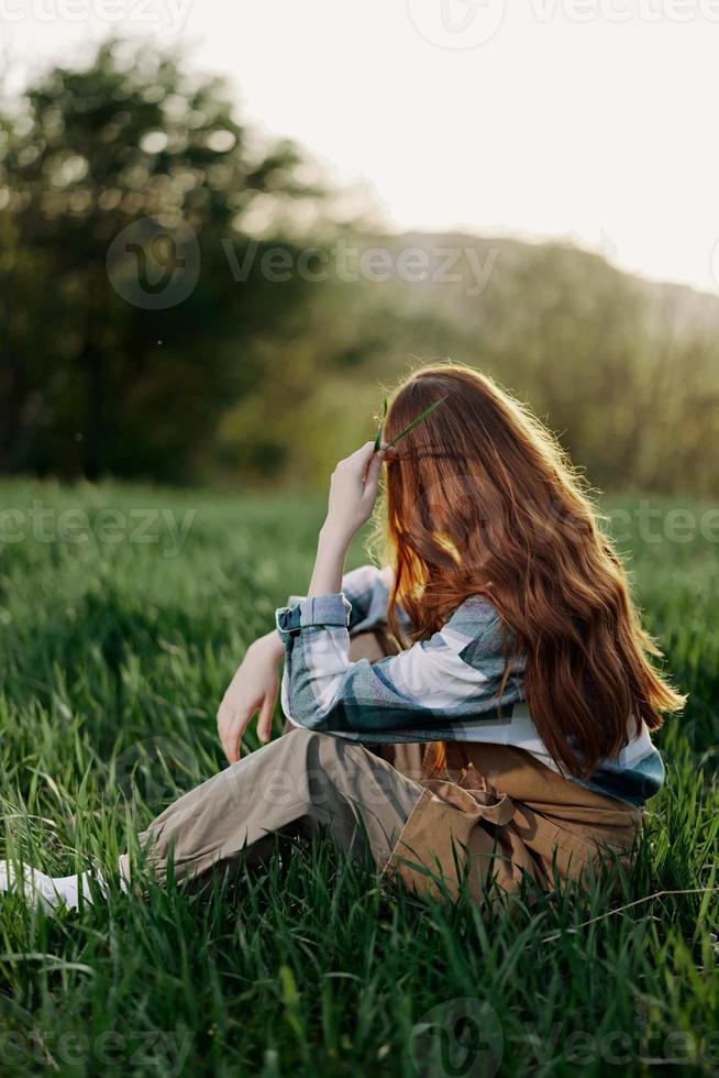 A woman sits on the green grass in a park with her back to the camera and relaxes in nature in the summer sunset evening light photo