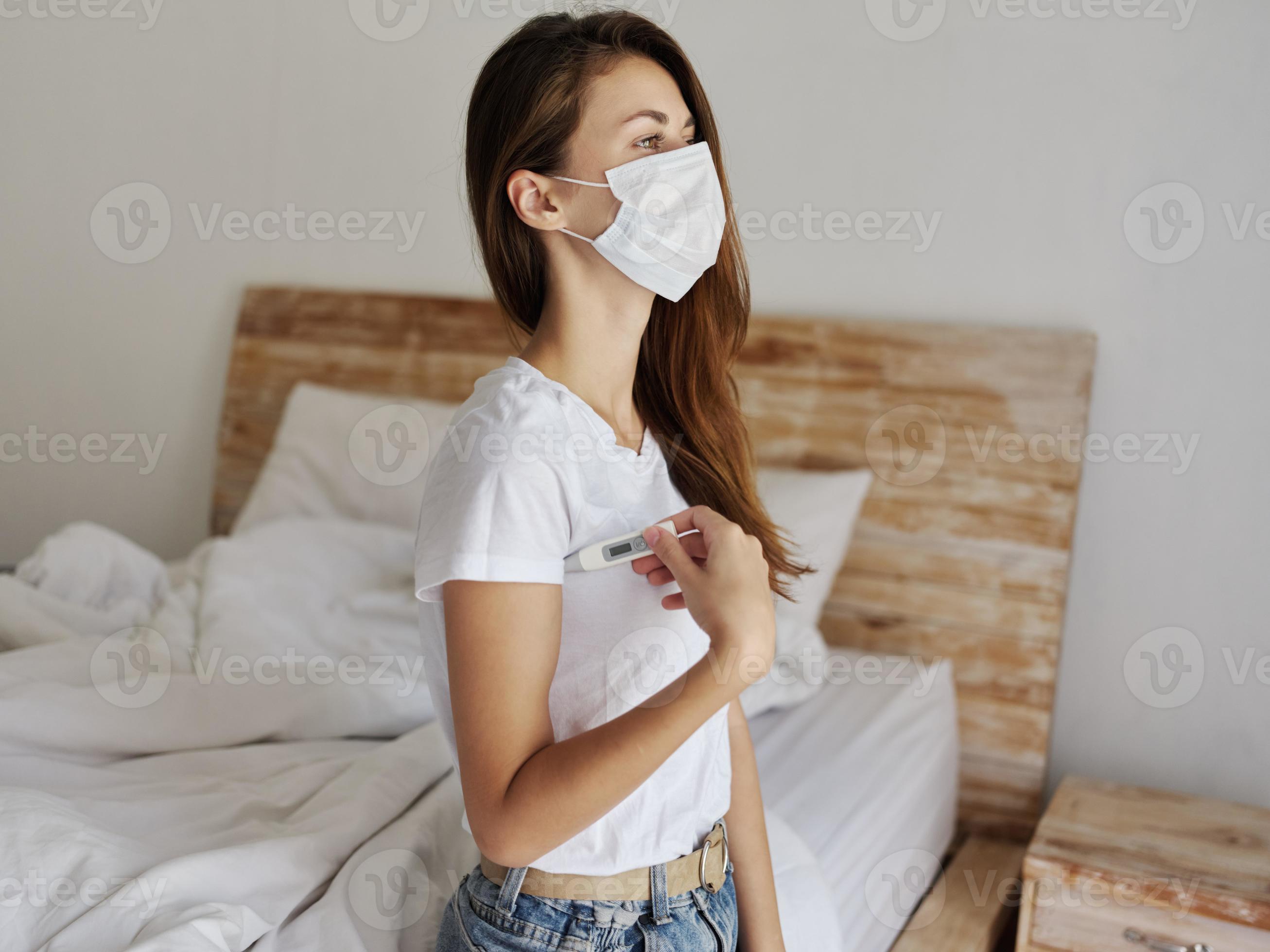 woman with thermometer under her arm medical mask sits on bed 22053888