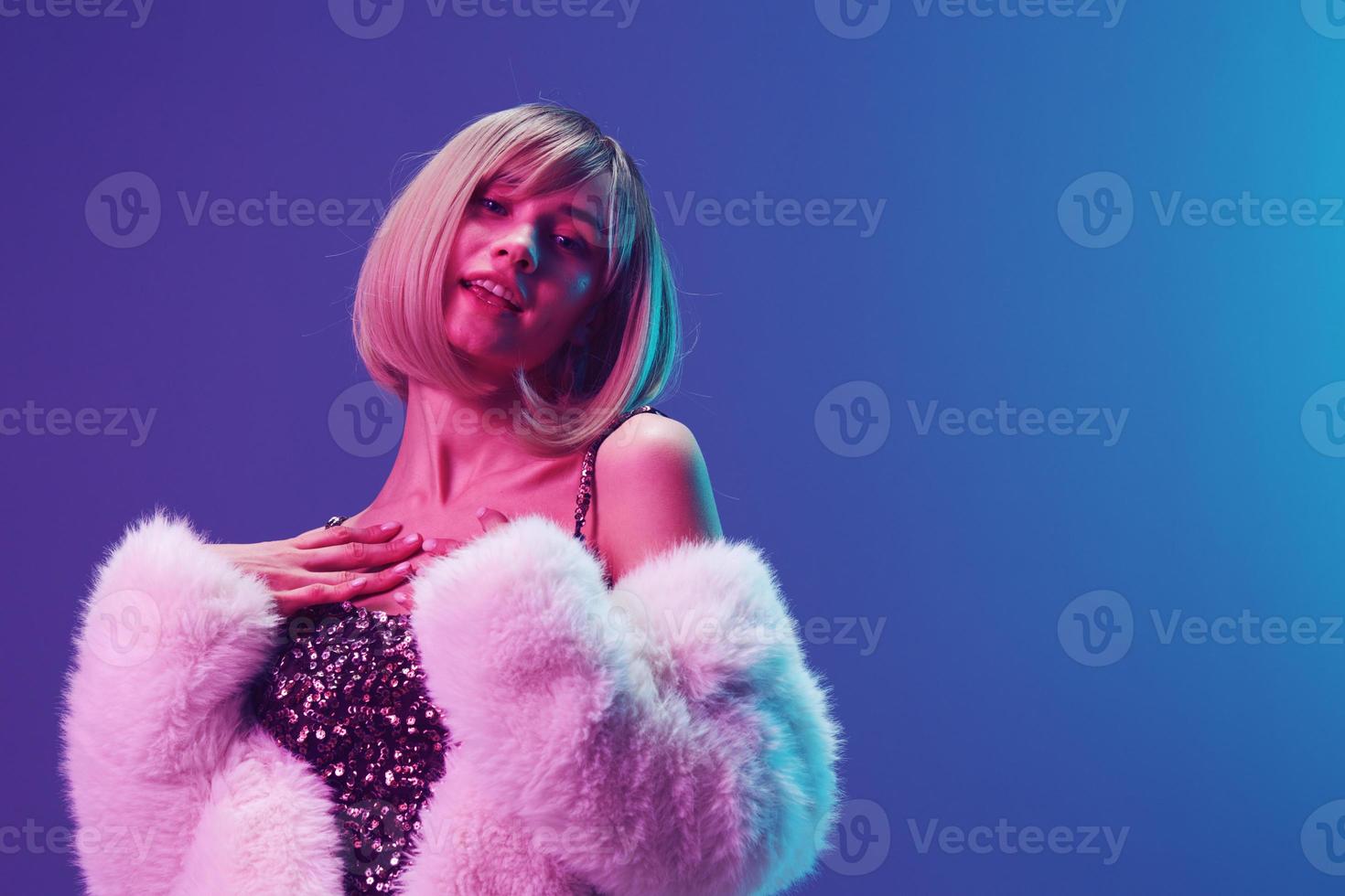 Enjoyed adorable blonde woman in fluffy fur coat sparkly dress hold hands on chest posing isolated in blue violet pink color light studio background. Neon party Fashion concept. Copy space Banner photo