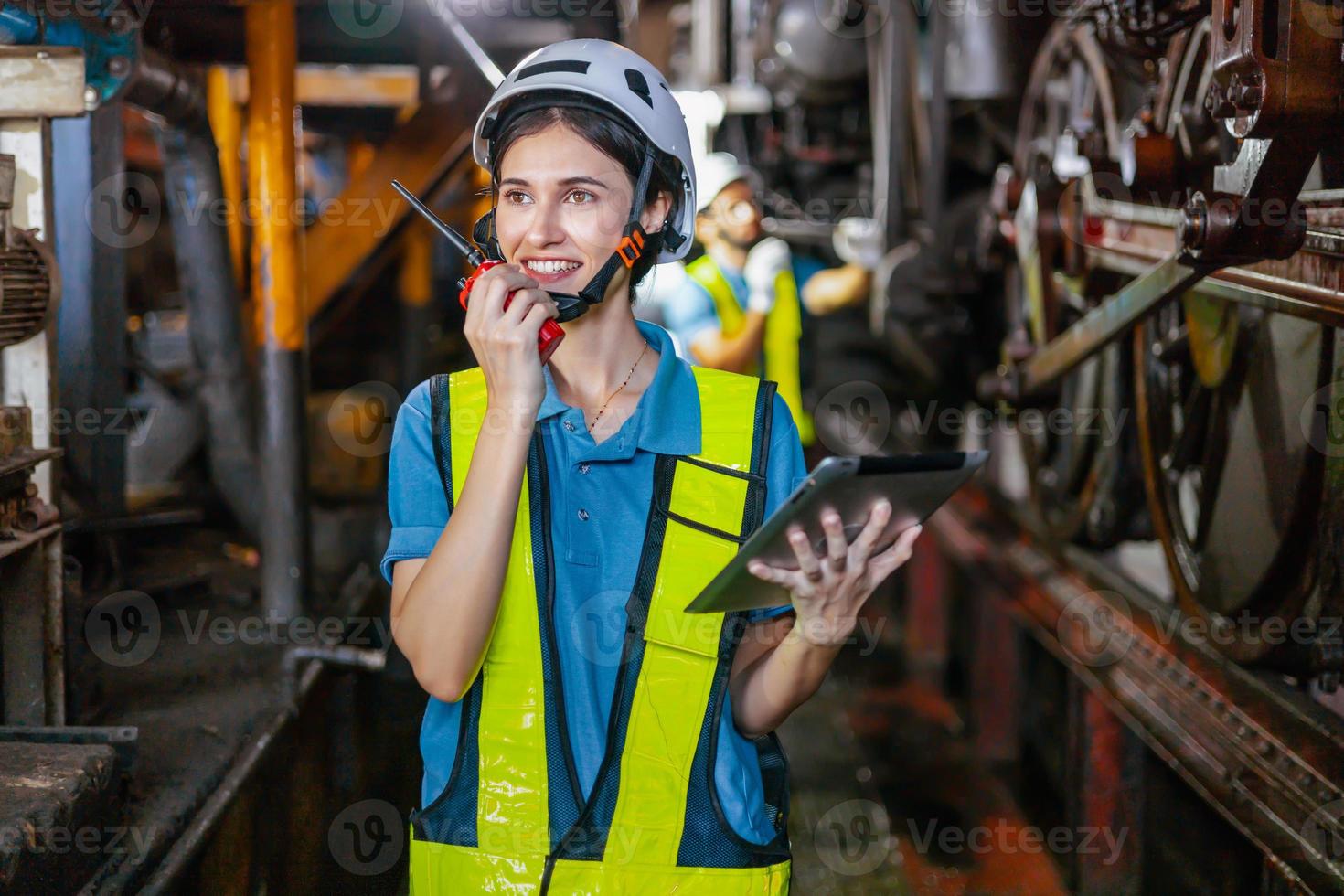 Beautiful woman engineer in factory talking on the radio wearing white helmet and work clothes and hand holding tablet at workplace photo