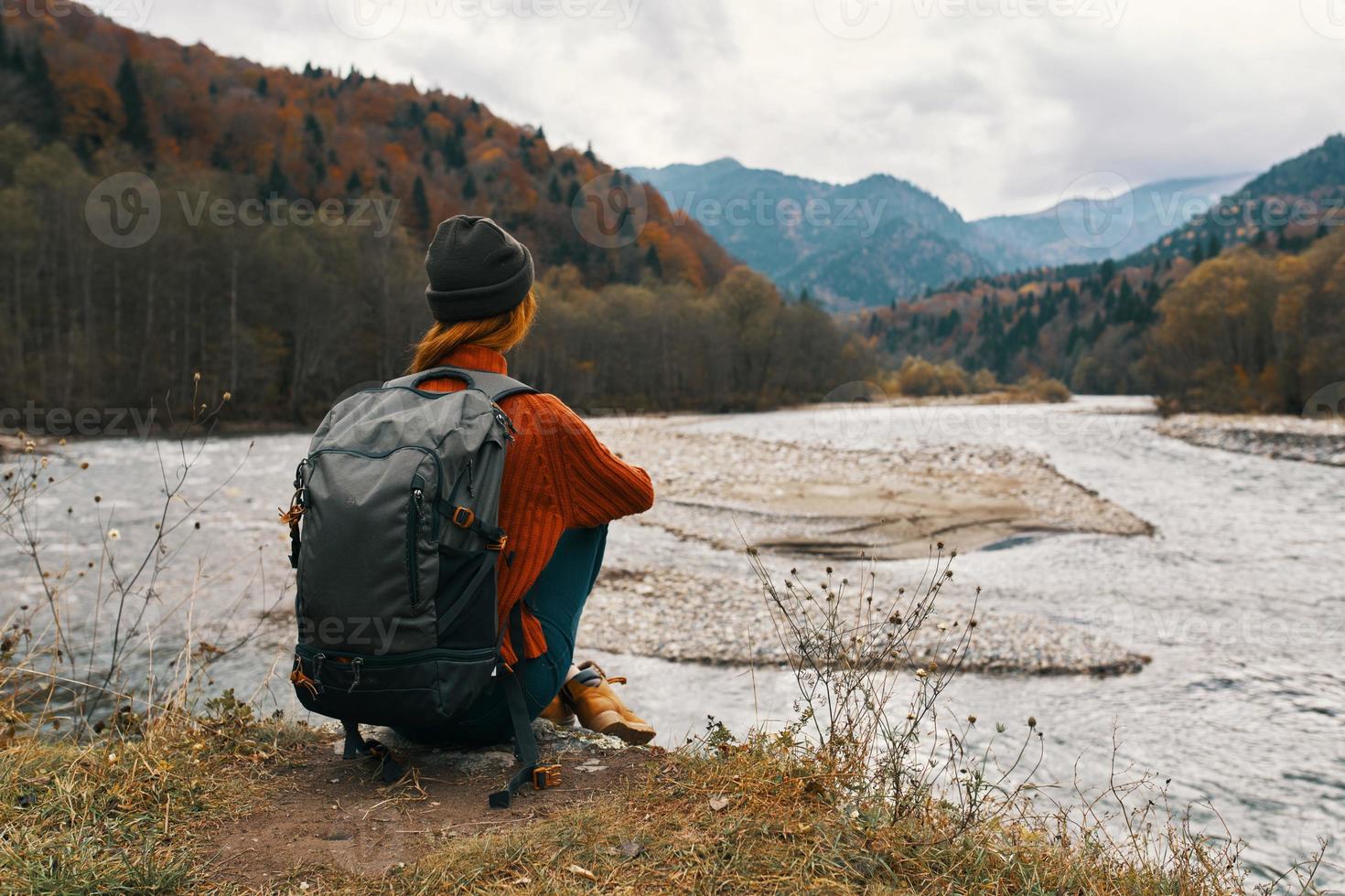 woman in the mountains in autumn with a backpack Sits on the river bank and looks at the high mountains photo