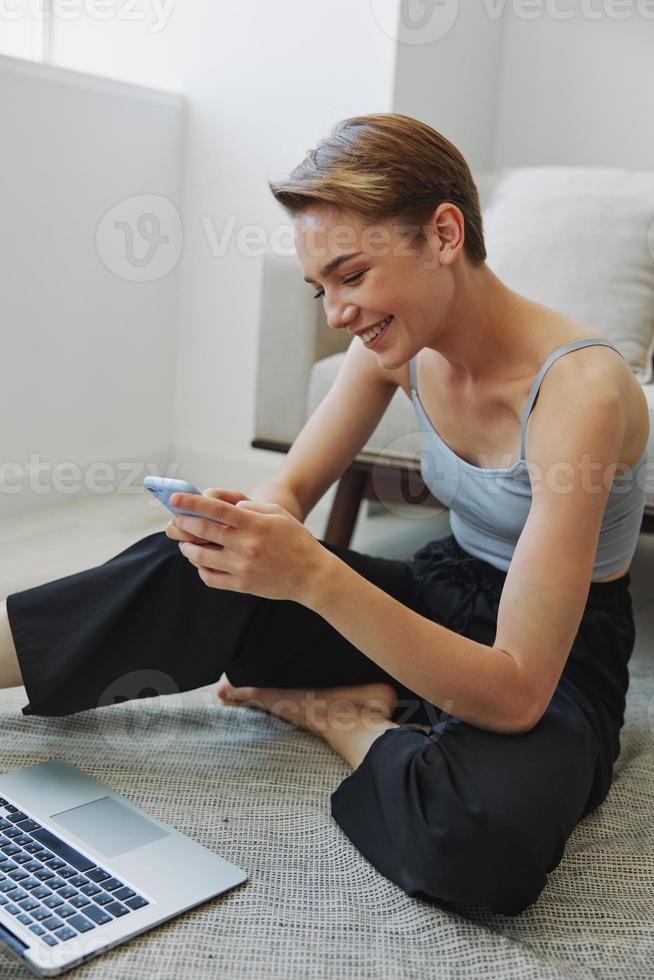 Freelance woman with laptop and phone works from home sitting on the floor in her home clothes with a short haircut, free copy space photo