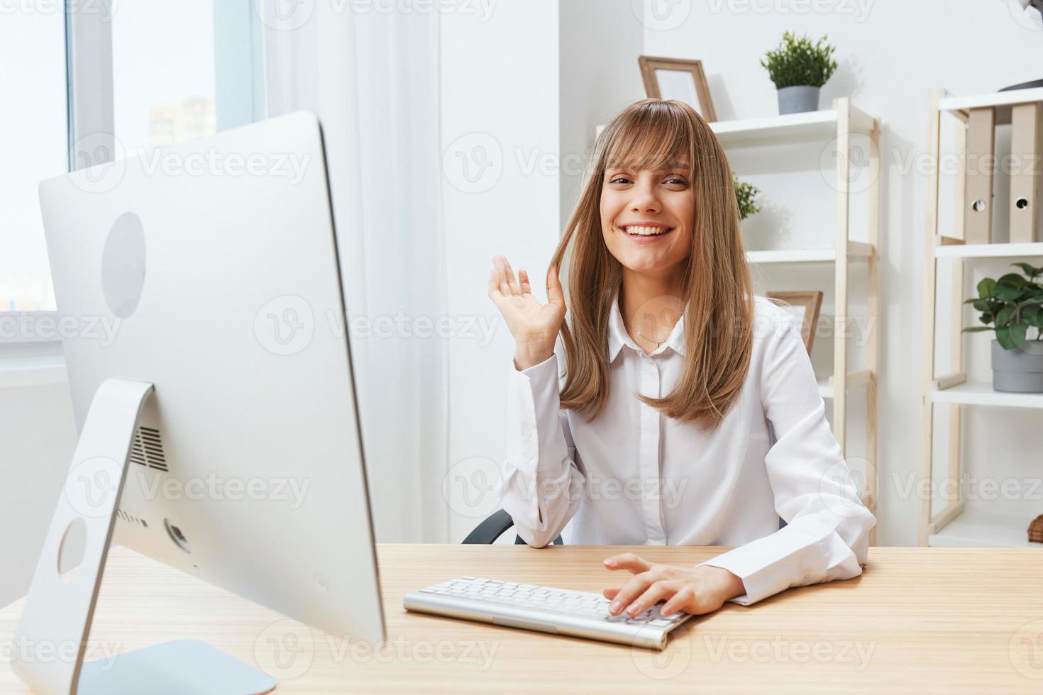 Smiling cheerful adorable blonde businesswoman worker freelancer look at camera raise hand up in light modern office. Happy employee work on computer online, enjoy successful business. Copy space photo