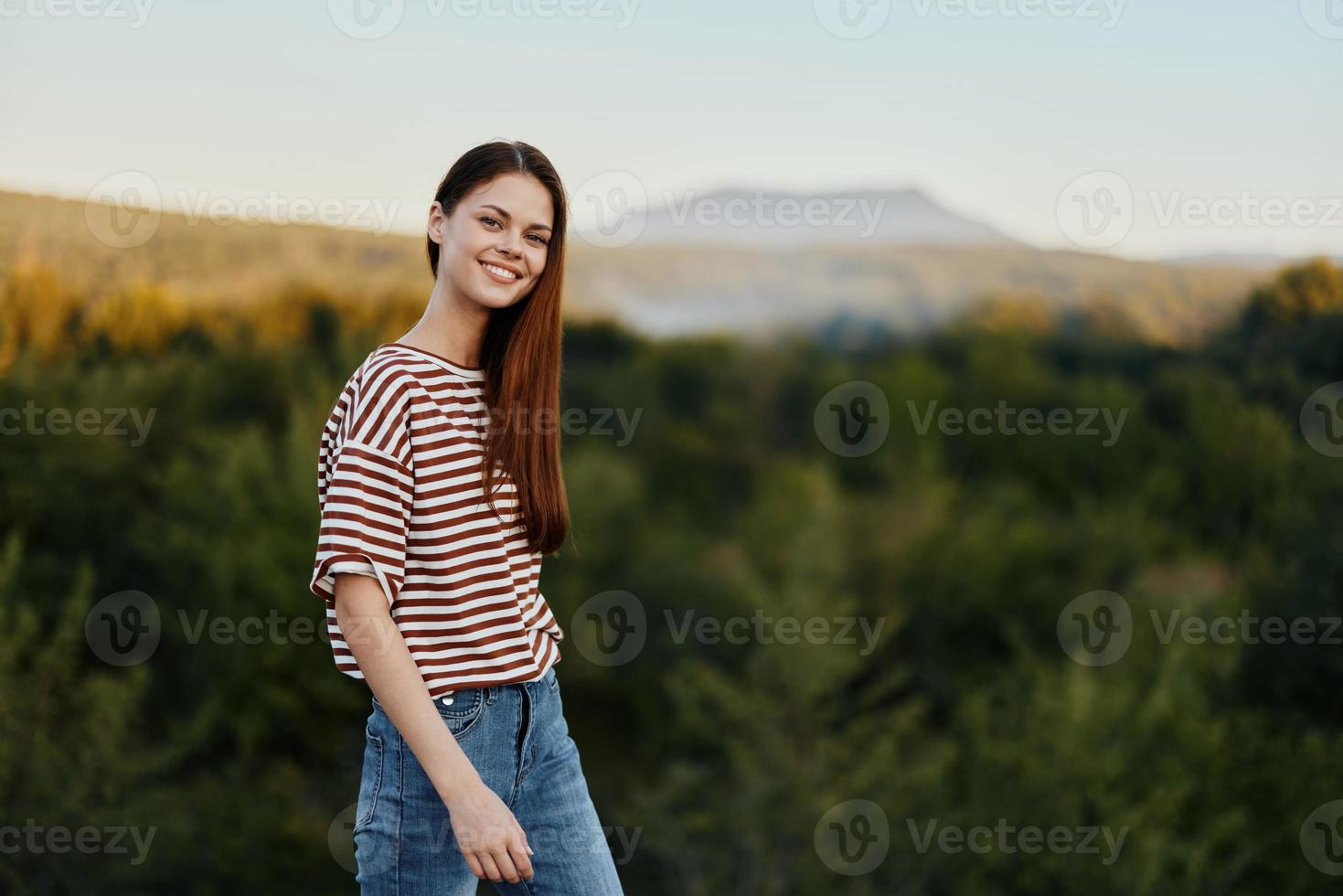 A young woman laughs and looks at the camera in simple clothes against the backdrop of a beautiful landscape of mountains and trees in autumn. Lifestyle on the move photo