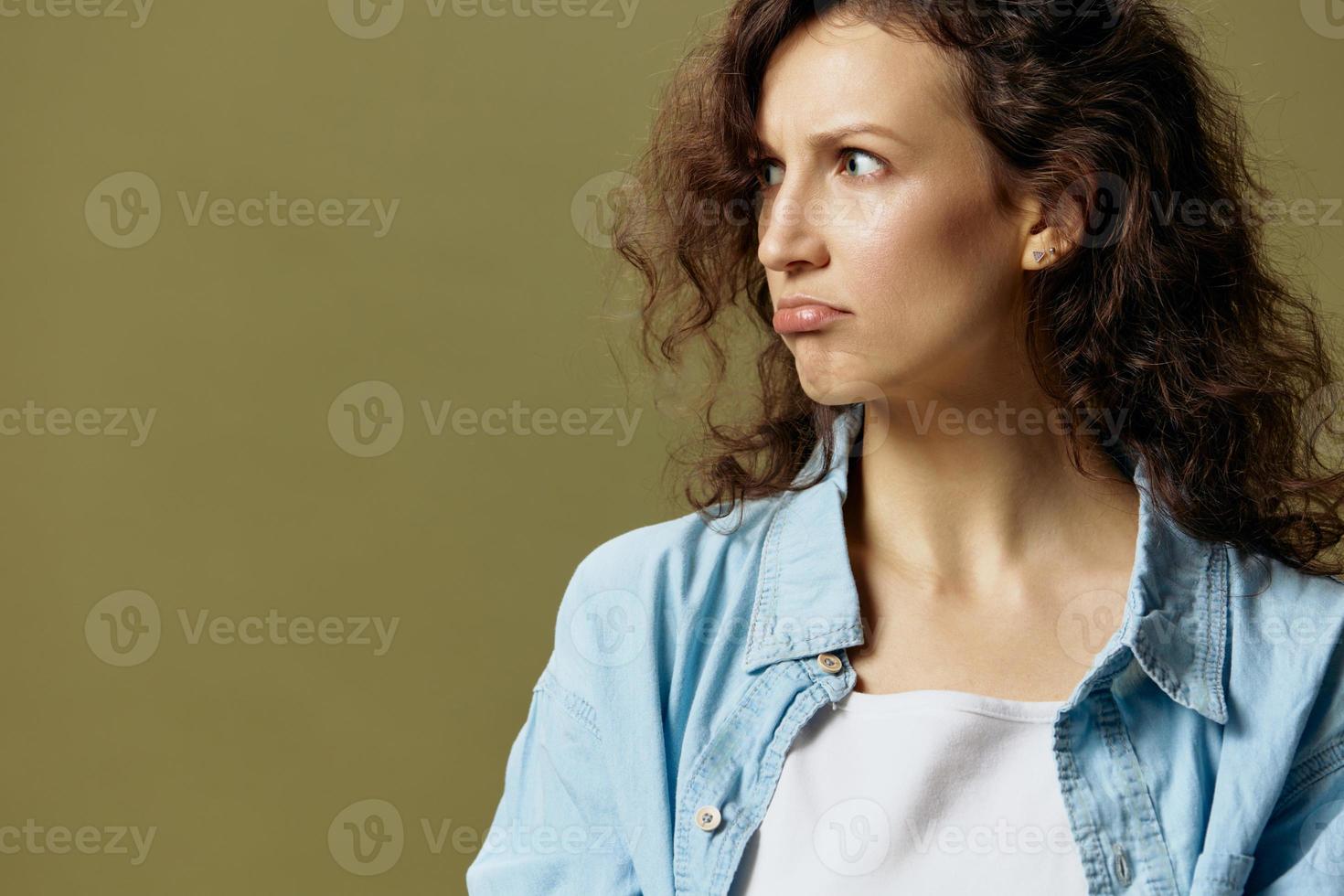 Upset unhappy curly beautiful female in jeans casual shirt frowns looks aside posing isolated on over olive green pastel background. Being Yourself. People Lifestyle emotions concept. Copy space photo