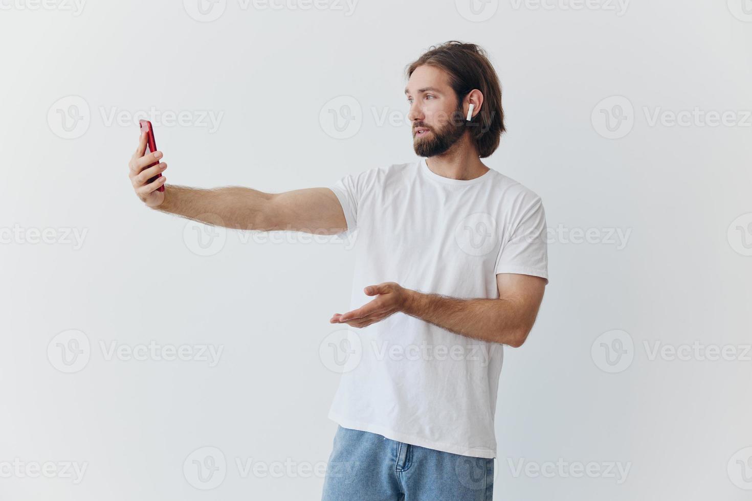 A man with a beard blogger in a white T-shirt with a phone and wireless headphones talking on an online video call against a white background photo