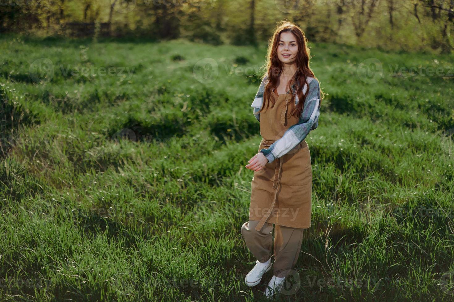 un mujer jardinero en un delantal soportes en un campo de verde césped al aire libre, sonriente en un verano tarde dentro un soleado puesta de sol después un días trabajo foto