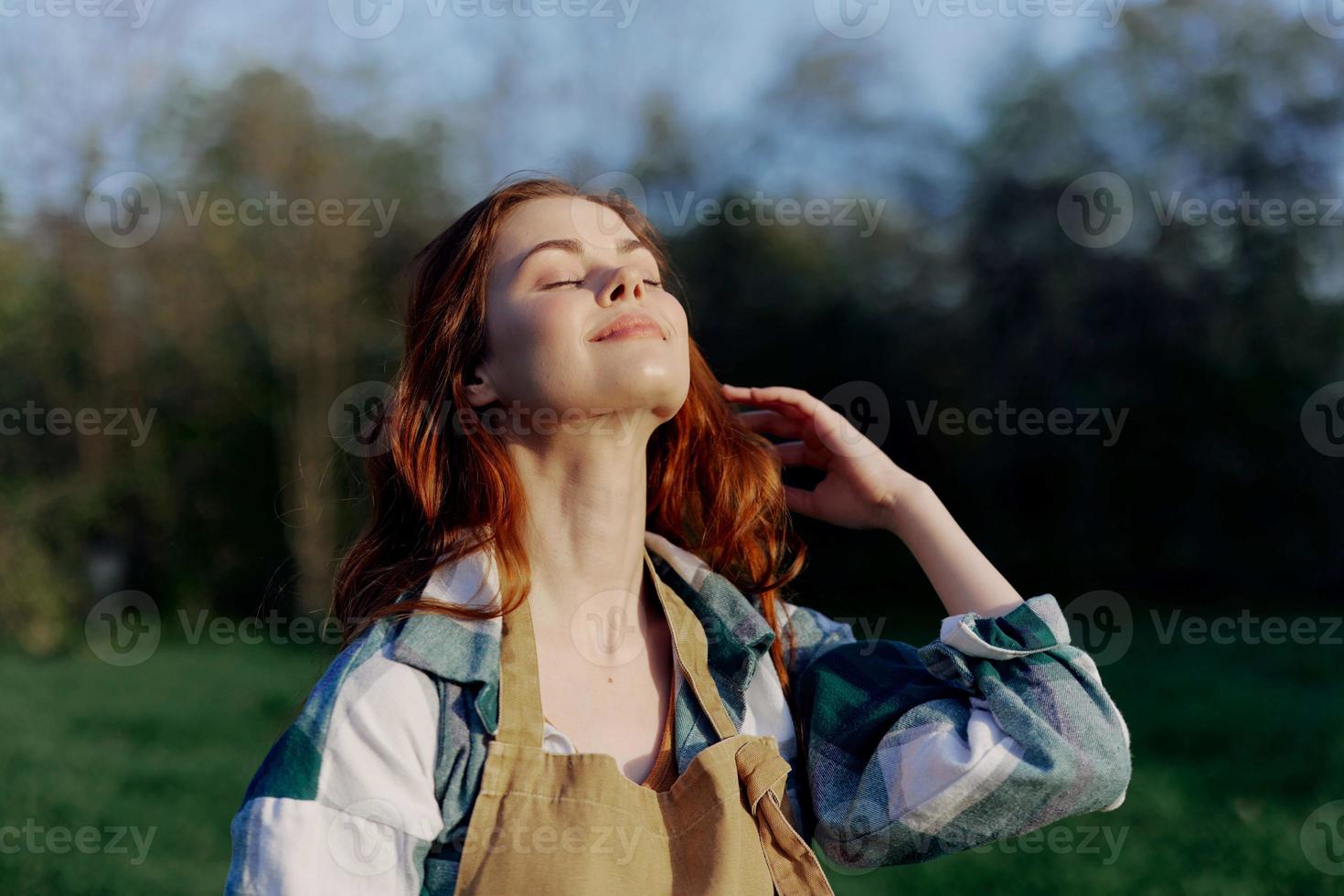 Portrait of a beautiful young woman with red hair in nature in a park in the summer, looking at the sunset against a background of green grass and trees photo